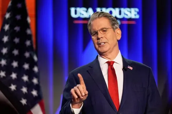 رجل يرتدي بدلة رسمية وقميص أبيض، يتحدث بجدية أمام لافتة تحمل عبارة "USA House" في منتدى دافوس الاقتصادي، مع العلم الأمريكي خلفه.