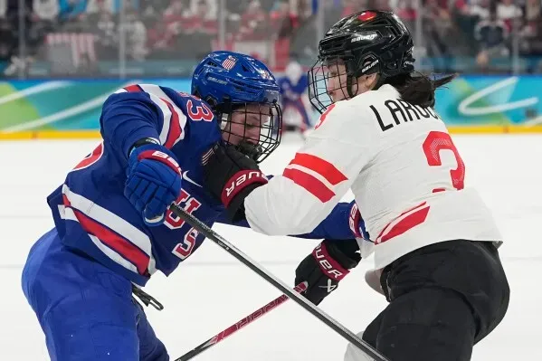 مباراة هوكي نسائية بين لاعبتين، حيث تتنافس لاعبة أمريكية ترتدي زيًا أزرق مع لاعبة كندية في زي أبيض، وسط أجواء حماسية.