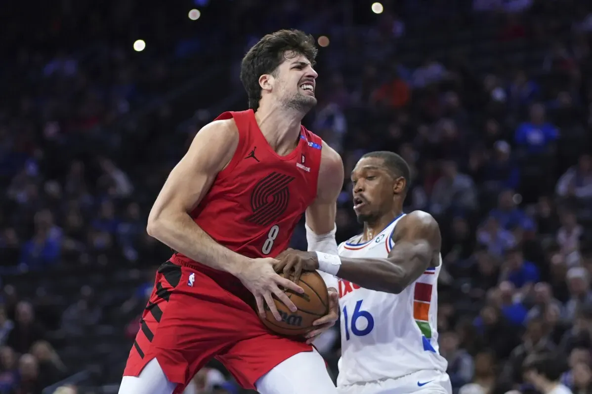 لاعب من بورتلاند تريل بليزرز يتنافس على الكرة مع لاعب من فيلادلفيا 76ers خلال مباراة كرة سلة، مع خلفية جماهيرية.