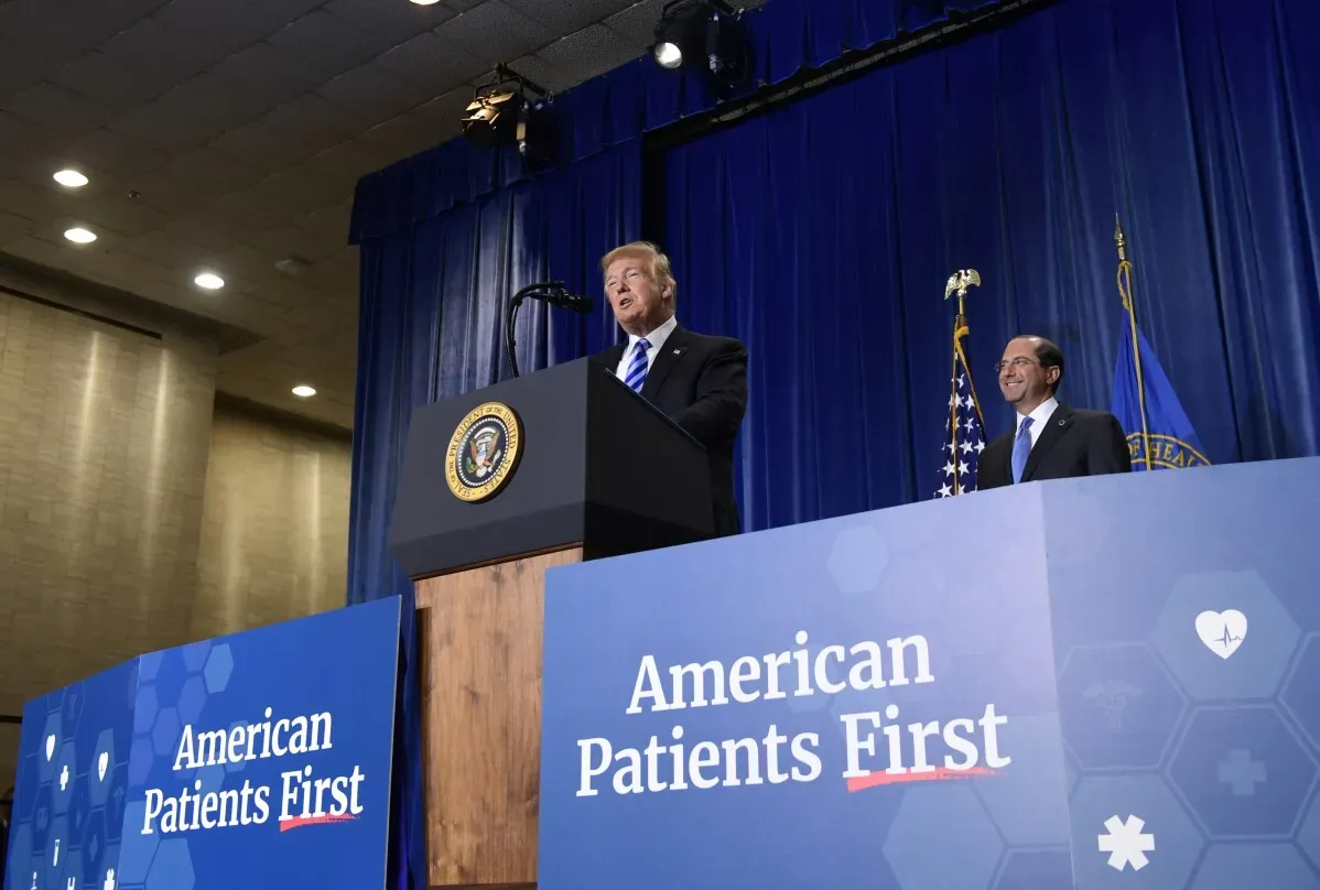 دونالد ترامب يتحدث في حدث صحي، بجانب لوجو "American Patients First"، مع وجود أعلام خلفية.