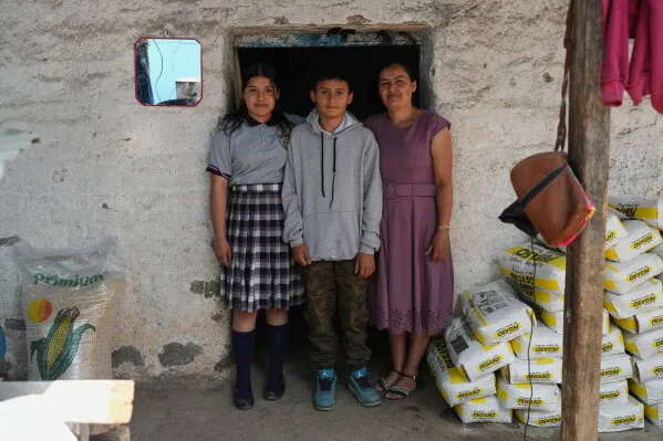 عائلة غواتيمالية تتكون من أم وطفلين يقفون أمام مدخل منزل بسيط، مع أكياس مواد غذائية خلفهم، يعكسون معاناة الأسر المفرقة بسبب سياسات الهجرة.
