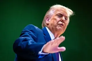 تظهر الصورة دونالد ترامب أثناء حديثه، مع تعبير جاد، في خلفية خضراء، مما يعكس سياسته المثيرة للجدل بشأن المساعدات الخارجية.