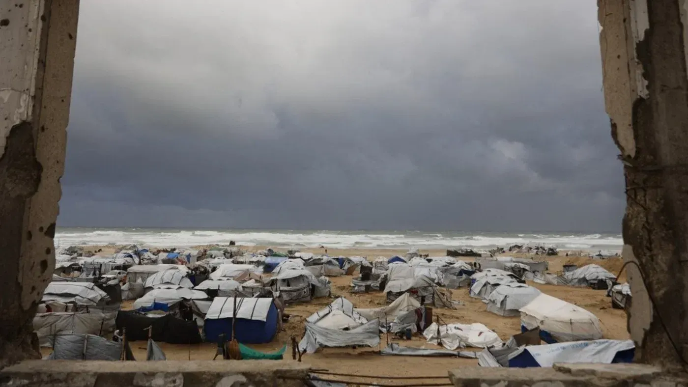 منظر من نافذة مدمرة يظهر مخيمات اللاجئين على شاطئ غزة تحت سماء غائمة، مما يعكس الوضع الإنساني الصعب في المنطقة.