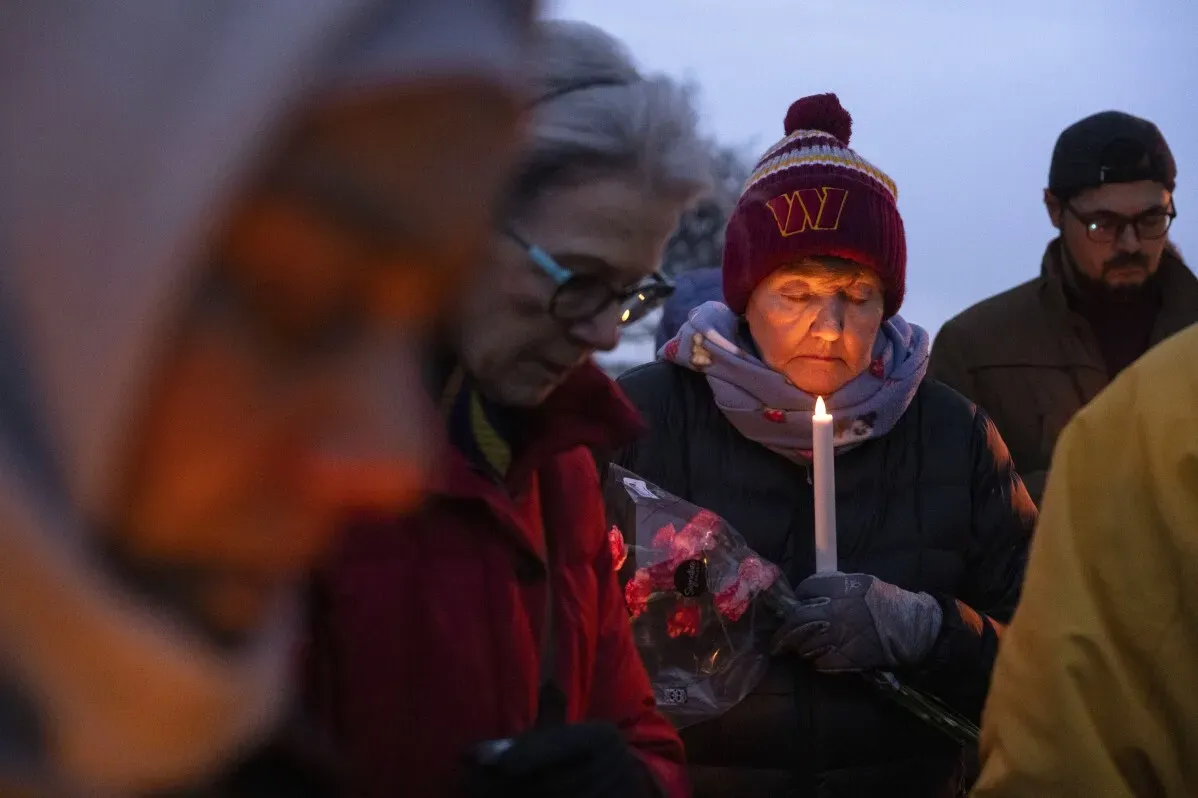 مجموعة من الأشخاص يشاركون في vigil تأبيني، يحمل أحدهم شمعة وزهور، مع تعبيرات حزينة في جو مظلم.