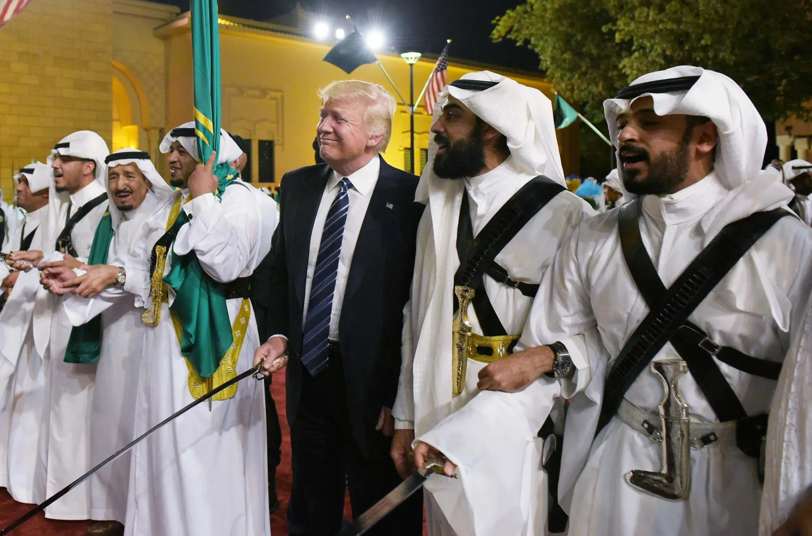ترامب يتفاعل مع مجموعة من الرجال يرتدون الزي التقليدي في حدث ثقافي، مع خلفية من الأعلام الأمريكية والسعودية.
