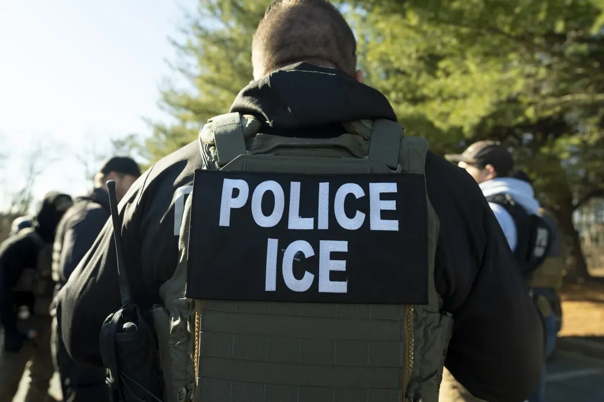 رتداء ضابط شرطة سترة تحمل كلمة "شرطة ICE"، مع خلفية تضم عناصر أمنية أخرى، خلال عملية تتعلق بالهجرة.