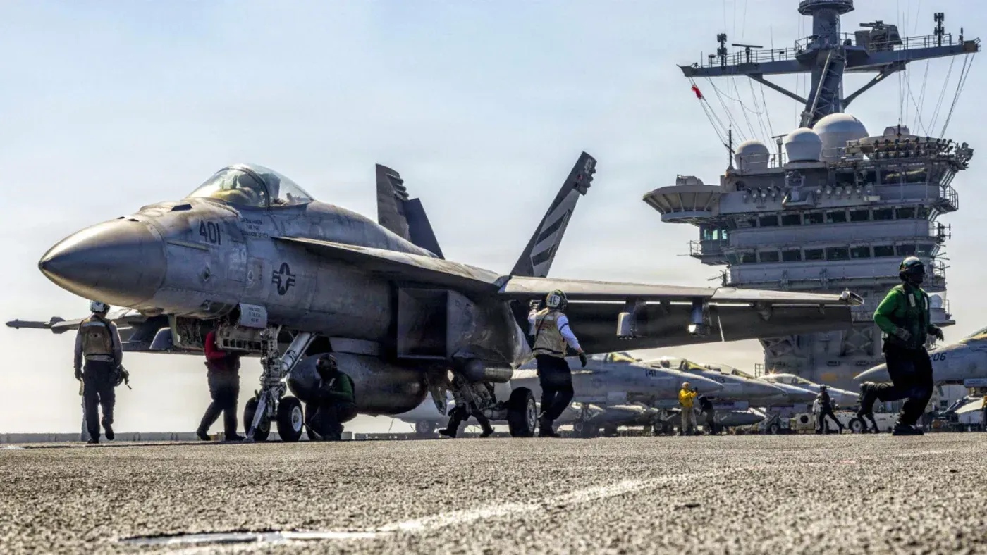 طائرة حربية من طراز F/A-18 هورنت على متن حاملة طائرات، مع أفراد الطاقم يتجهون نحو الطائرة، مما يعكس النشاط العسكري الأمريكي في المنطقة.