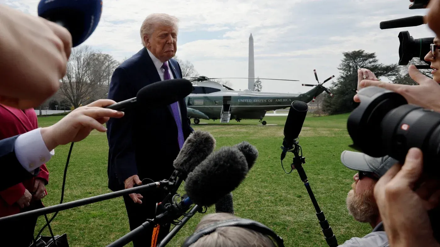 ترامب يتحدث للصحفيين خارج البيت الأبيض بينما تحيط به الميكروفونات، مع وجود مروحية في الخلفية ونصب واشنطن في المسافة.