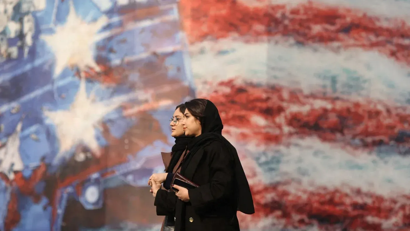 امرأتان ترتديان الحجاب تسيران معًا أمام جدارية تحمل ألوان العلم الأمريكي، مما يعكس التوترات السياسية بين إيران والولايات المتحدة.