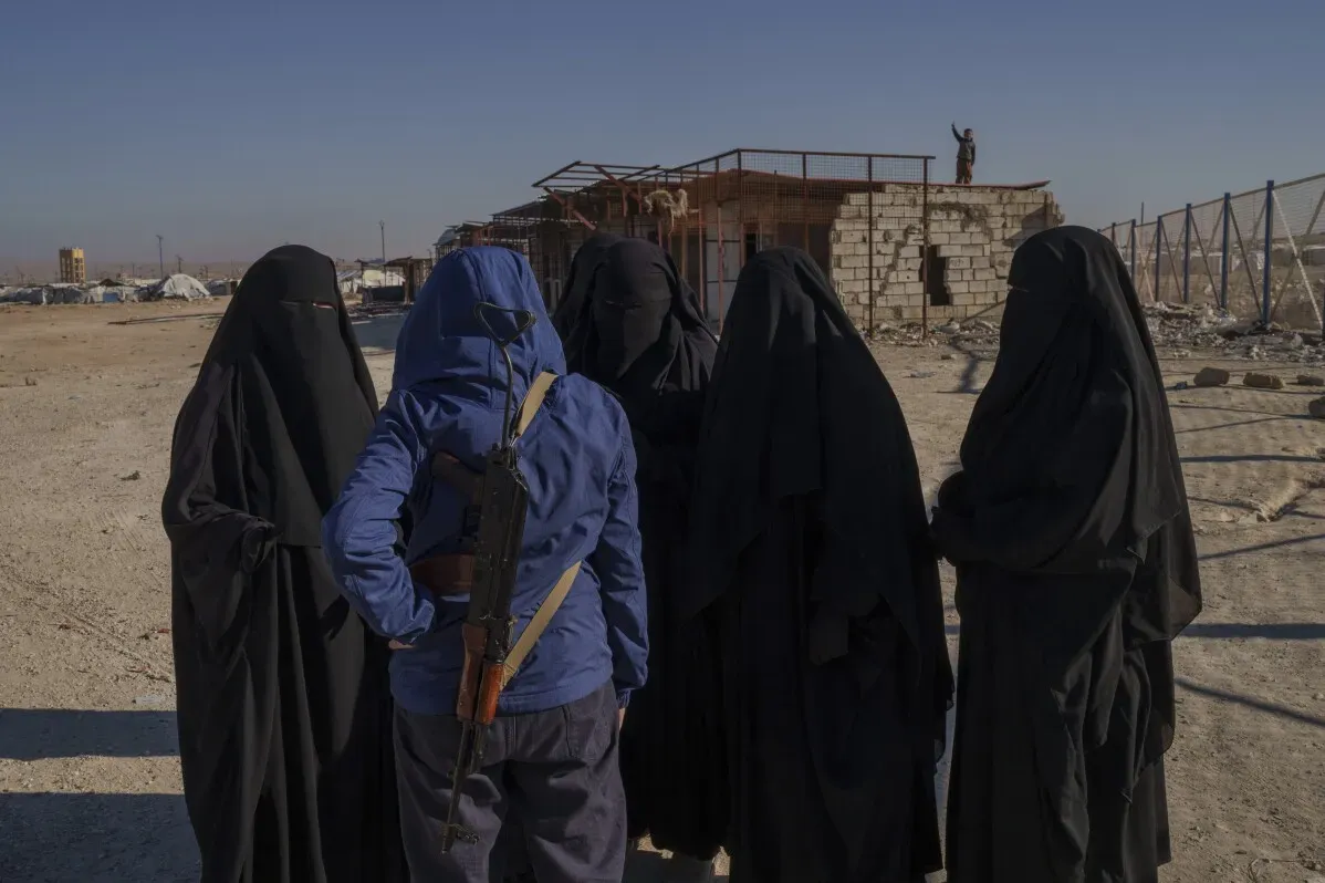 نساء يرتدين العباءات السوداء يتحدثن مع امرأة ترتدي ملابس زرقاء وتحمل سلاحًا، في مخيم الهول بسوريا، حيث تتزايد الأوضاع الإنسانية سوءًا.