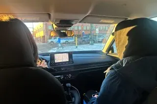 مشهد من داخل سيارة يظهر شخصين يجلسان في المقعد الأمامي، مع التركيز على شاشة نظام الملاحة، في منطقة حضرية بمينيابوليس.