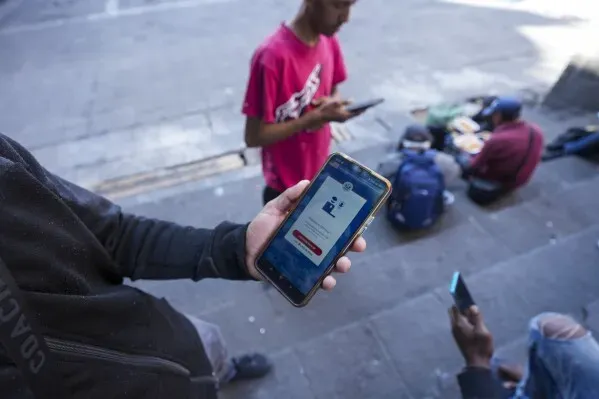 شخص يحمل هاتفًا ذكيًا يظهر تطبيق CBP One، بينما يقف آخرون في الخلفية، مما يعكس التحديات التي يواجهها المهاجرون في الولايات المتحدة.