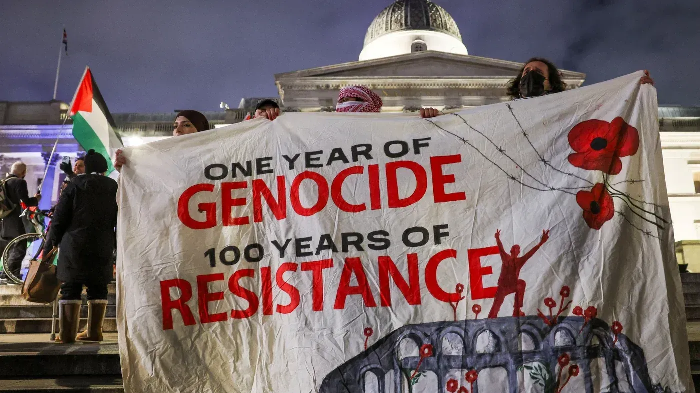 لافتة تحمل عبارة "عام من الإبادة، 100 عام من المقاومة" خلال احتجاج لدعم حقوق الفلسطينيين، مع علم فلسطيني في الخلفية.