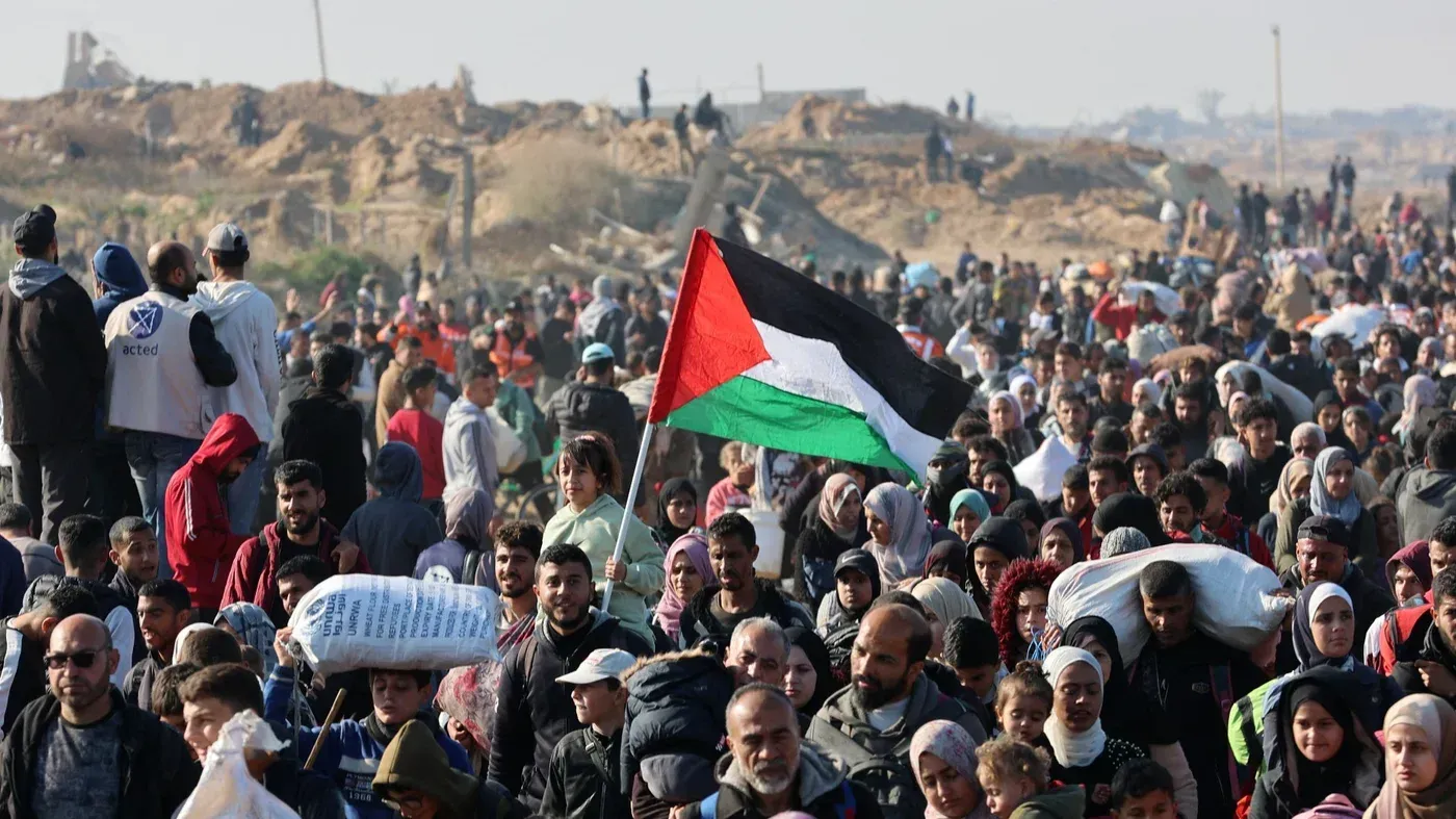 جموع من الفلسطينيين في منطقة غزة يحملون الأعلام ويعبرون عن معاناتهم، وسط أكوام من الحطام، في سياق أزمة النزوح.