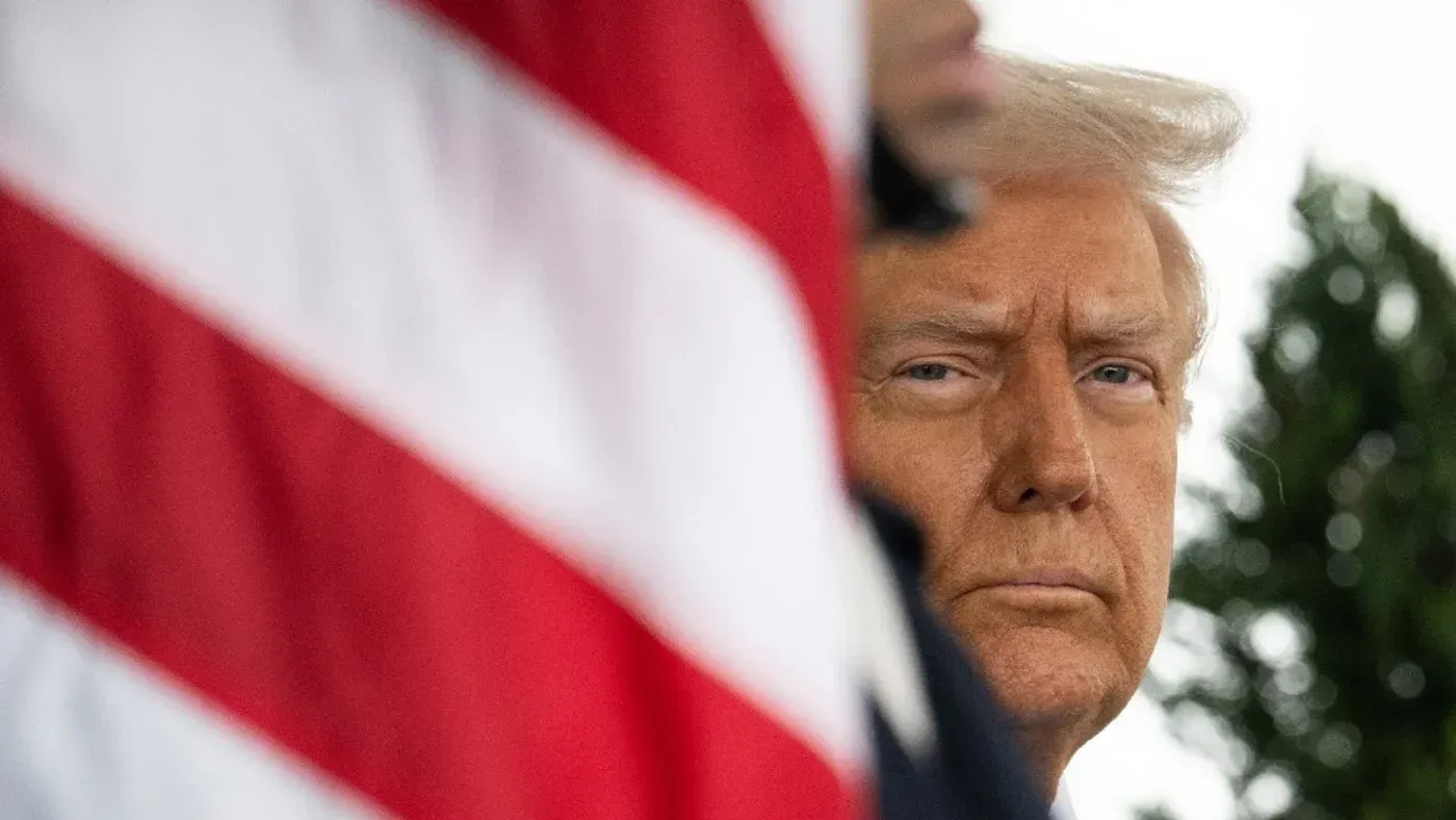 صورة لدونالد ترامب يظهر فيها بملامح جادة، مع العلم الأمريكي خلفه، تعكس سياسته الاقتصادية تجاه إيران وتأثيرها على الطبقة الوسطى.