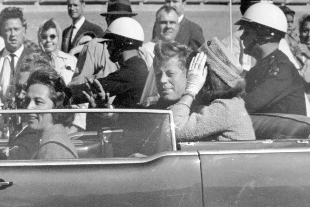 جون كينيدي وزوجته جاكلين في سيارة مكشوفة، محاطين بحضور جماهيري وضباط شرطة، أثناء زيارتهما لدالاس عام 1963.
