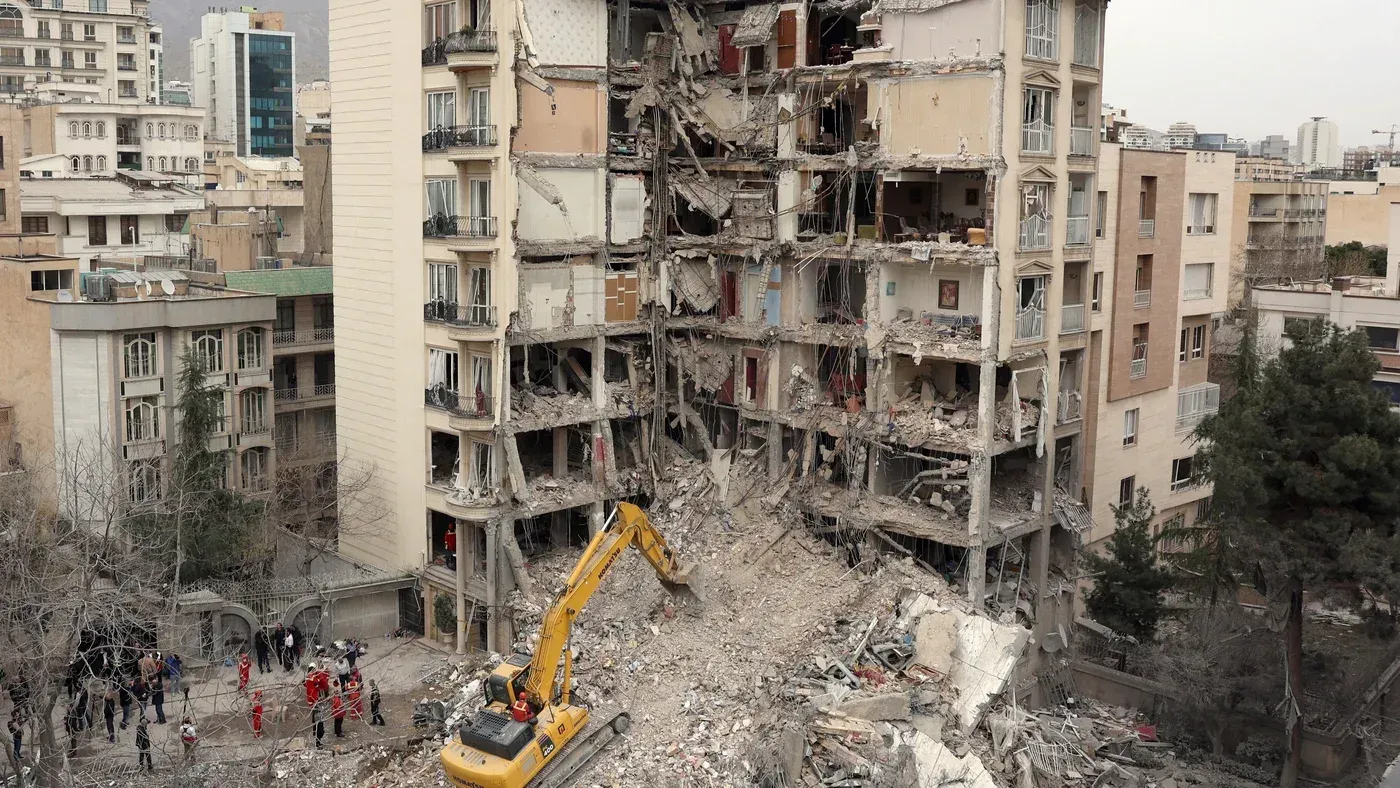 مبنى سكني مدمّر جزئيًا في طهران، مع وجود جرافة وعمال إنقاذ في الموقع، يعكس آثار الصراع والتوترات الجيوسياسية في المنطقة.