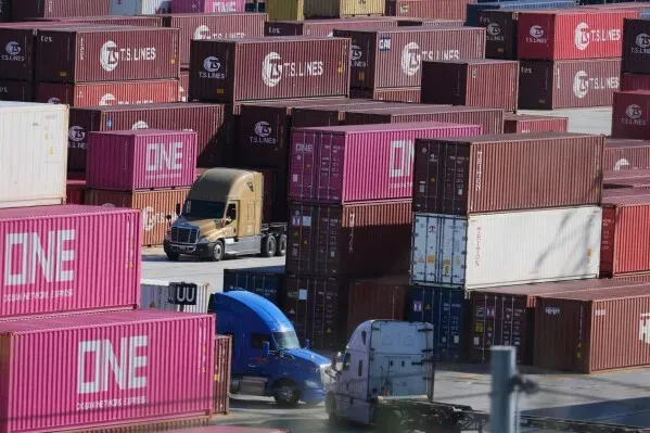 شاحنات تسير بين حاويات الشحن الملونة في ميناء، تعكس تأثير التعريفات الجمركية على التجارة العالمية والاقتصاد الأمريكي.