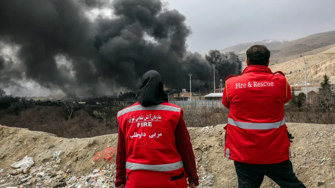 رجال إطفاء يرتدون زيًا أحمر يقفون أمام سحابة من الدخان الأسود الكثيف، في مشهد يعكس حالة الطوارئ بعد الهجوم على منشأة.