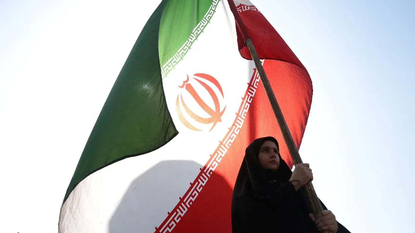 امرأة ترتدي الحجاب تحمل علم إيران، مما يعكس الأجواء الوطنية والتوترات الإقليمية في سياق الحرب على إيران وتأثيراتها.