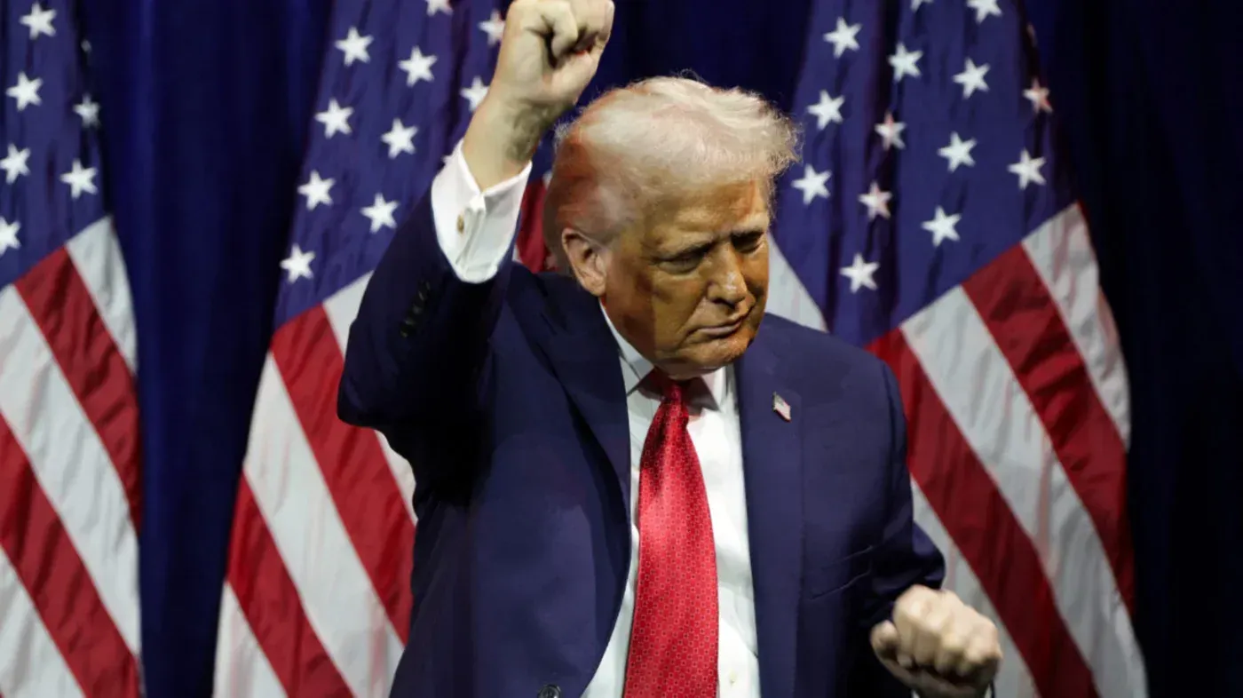 ترامب يرفع يده في خطاب، مع أعلام أمريكية خلفه، معبراً عن طموحاته في السيطرة على الثروات النفطية لفنزويلا.