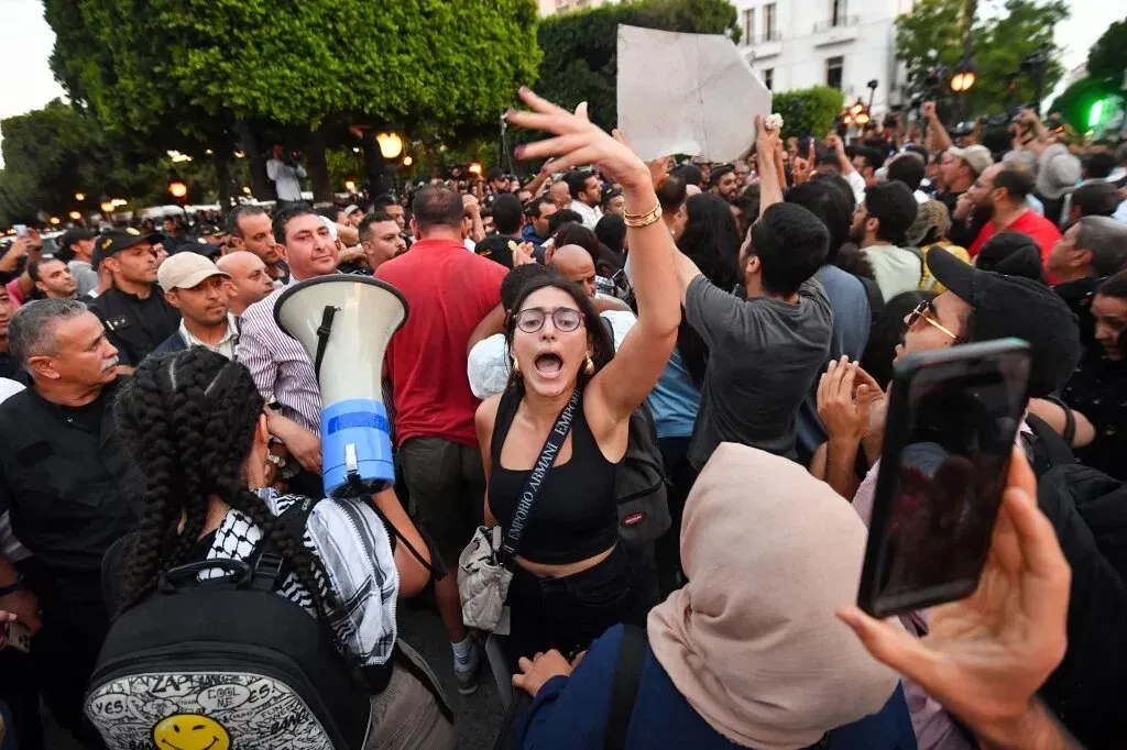 احتجاجات في تونس، حيث تتجمع حشود كبيرة، مع امرأة تحمل مكبر صوت وتعبّر عن مشاعر الغضب والمطالبة بالتغيير الاجتماعي.