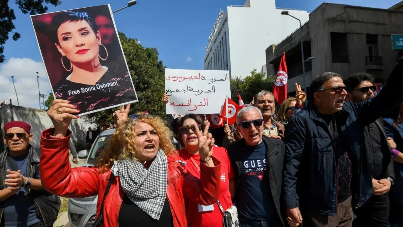 محتجون في تونس يرفعون لافتات تطالب بالإفراج عن الصحفية سونيا دحماني، مع تواجد أعلام تونسية في الخلفية، تعبيراً عن قمع الحكومة.