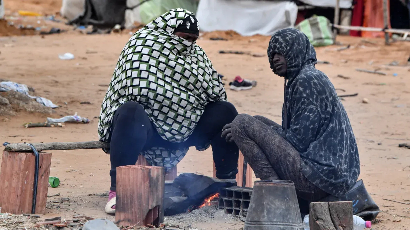 امرأتان تجلسان بالقرب من نار في مخيم للاجئين، يرتديان ملابس ثقيلة، تعكسان ظروف الحياة الصعبة للمهاجرين في تونس.