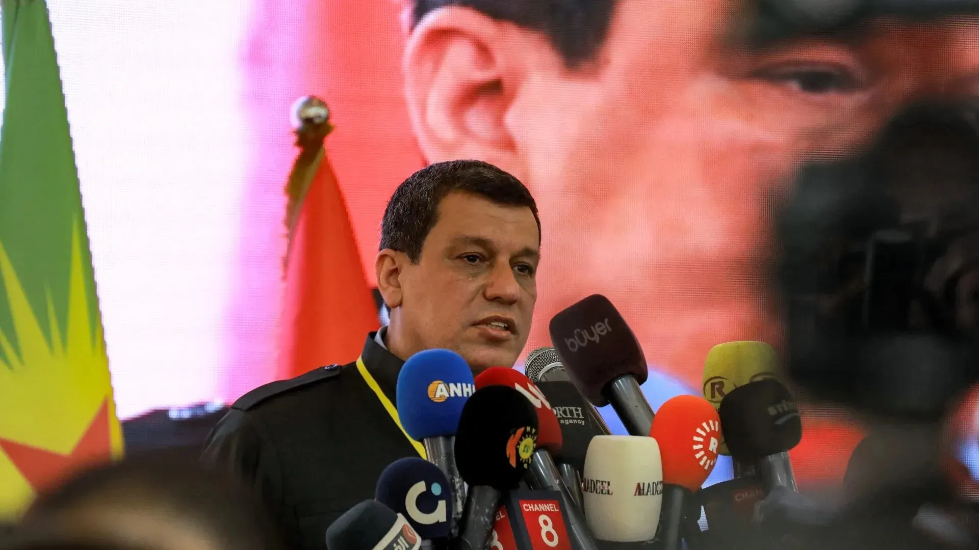 تظهر الصورة قائد قوات سوريا الديمقراطية وهو يتحدث في مؤتمر صحفي، مع وجود أعلام خلفه وميكروفونات متعددة، مما يعكس أهمية الحوار حول مستقبل سوريا.