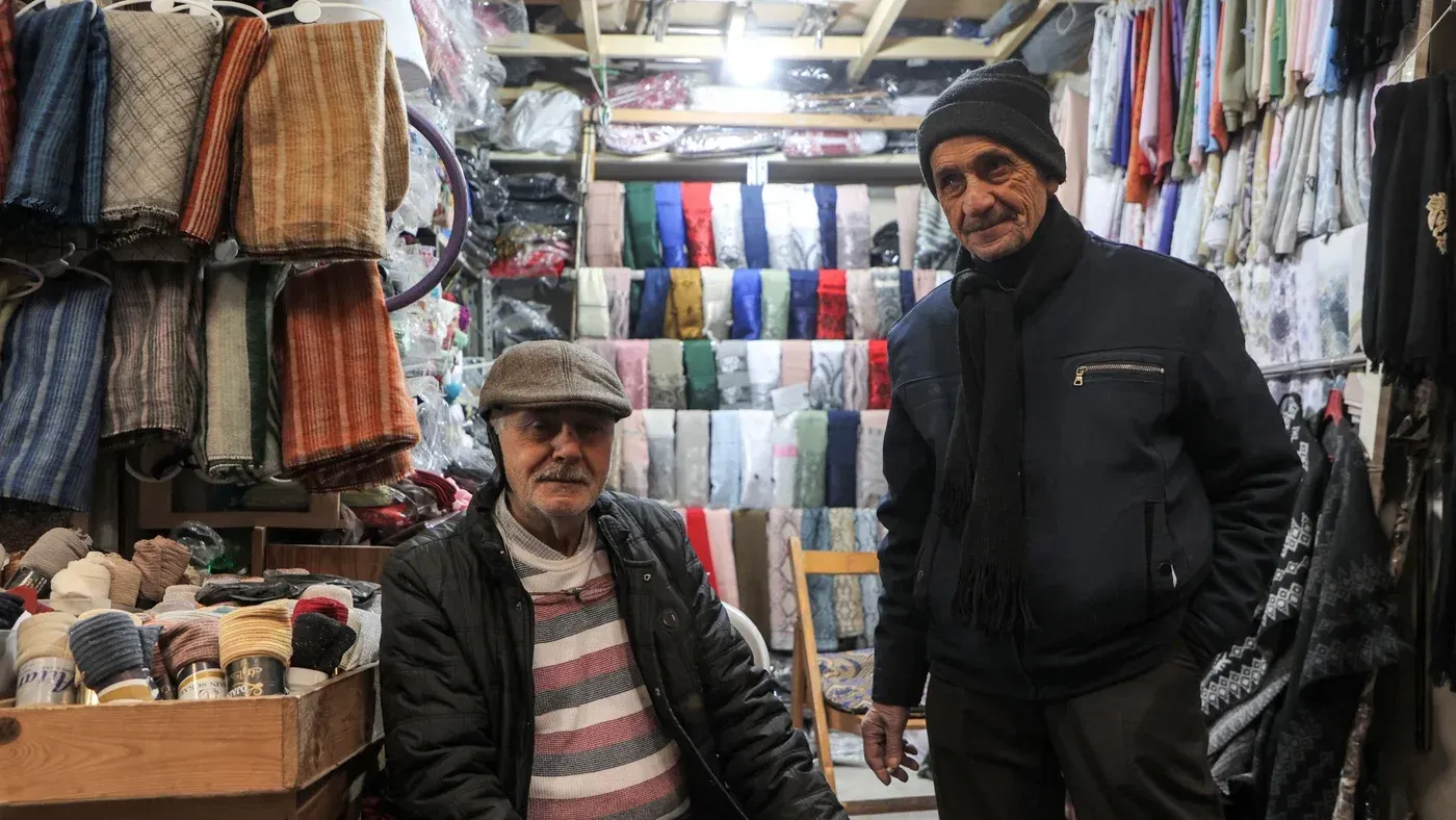 رجلان يجلسان في متجر ملابس، محاطان بأقمشة ملونة ومختلفة، يعكسان العلاقات التجارية المتجددة بين تركيا وسوريا.