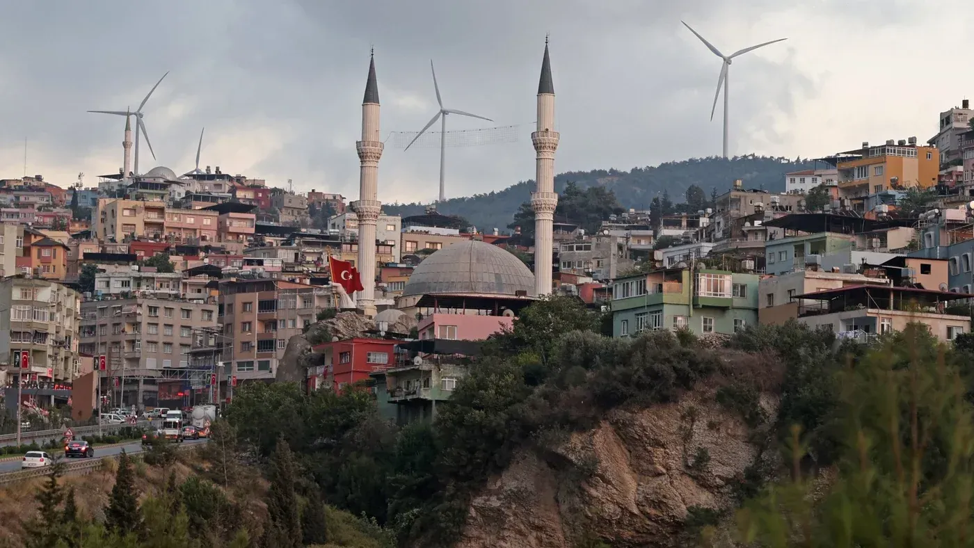 منظر لمدينة دورتيول في تركيا، يظهر مئذنة مسجد، مباني ملونة، وعلم تركي، مع توربينات رياح في الخلفية تعكس التقدم التكنولوجي.