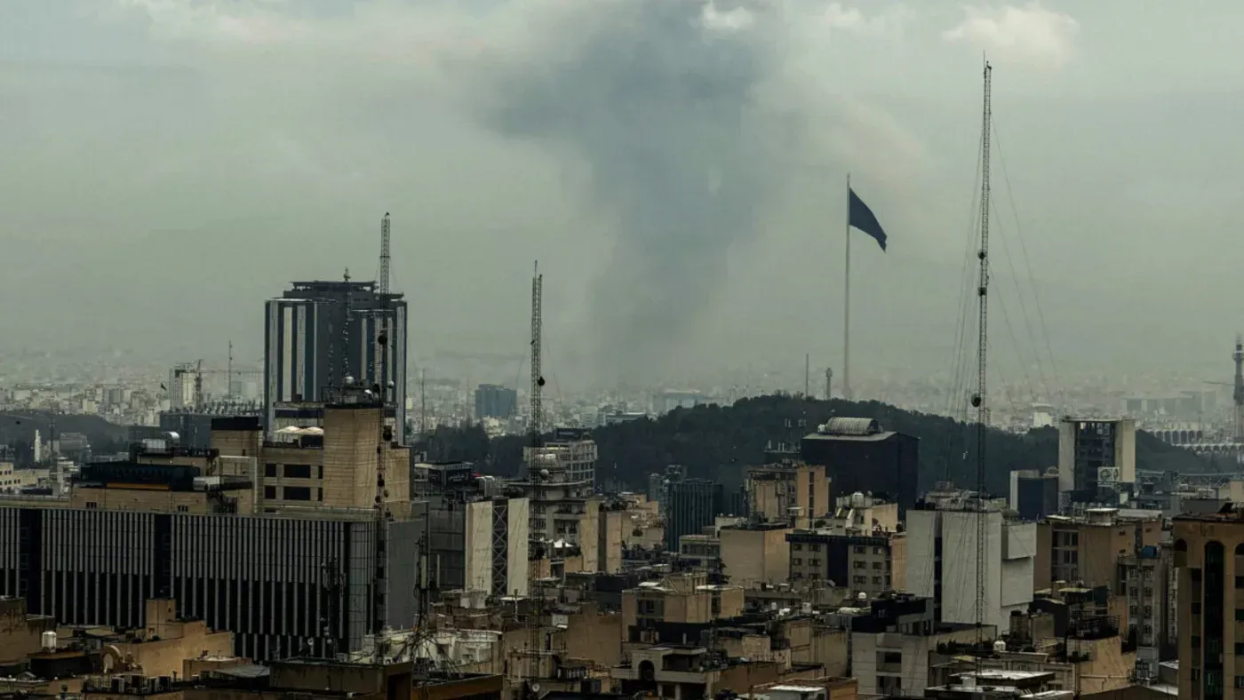 دخان يتصاعد من خلف مباني طهران، مع العلم الإيراني يرفرف في المقدمة، مما يعكس التوترات في المنطقة والجهود الدبلوماسية لإنهاء النزاع.