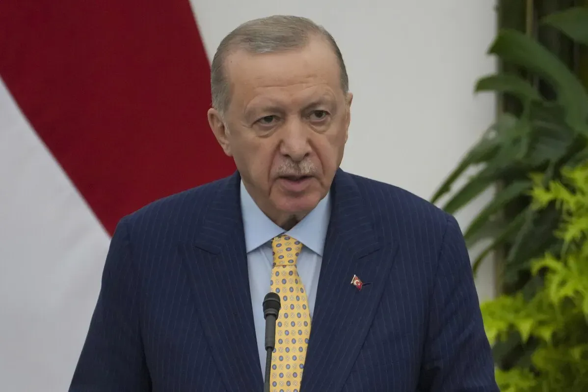 الرئيس التركي رجب طيب أردوغان يتحدث خلال مؤتمر، محاطًا بأعلام تركيا ونباتات، وسط توترات سياسية واقتصادية.