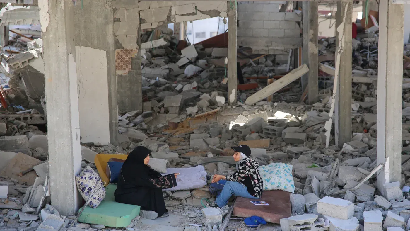 امرأتان تجلسان على وسائد بين أنقاض مستشفى مدمر، مع تواجد الحطام حولهما، مما يعكس آثار الدمار والخراب.