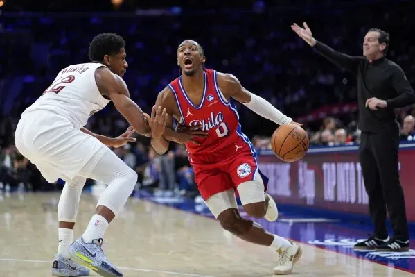 لاعب فريق 76ers تيريس ماكسي يتجاوز مدافع كليفلاند كافالييرز، مع وجود مدرب الفريق في الخلفية، خلال مباراة مثيرة.