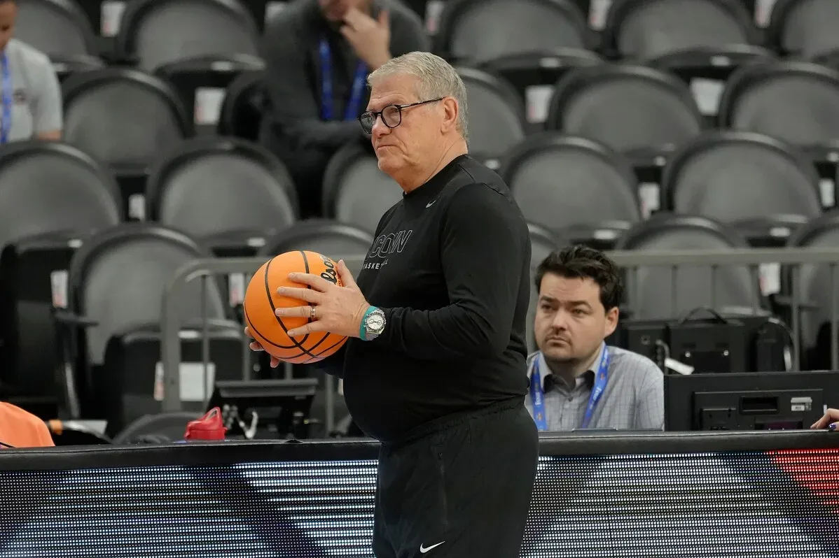 مدرب فريق يوكون يحمل كرة سلة، مستعداً للمباراة في بطولة NCAA للسيدات، بينما يجلس الصحفيون في الخلف.