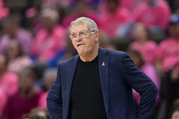 مدرب فريق كرة السلة النسائي في جامعة يوكُن، جينو أوريما، يتحدث خلال مباراة، بينما يرتدي الحضور ملابس وردية لدعم قضية معينة.