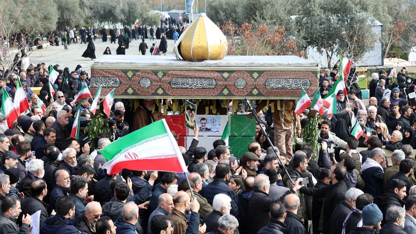 تجمع حشود من الناس في مناسبة وطنية في إيران، حاملين الأعلام الإيرانية، مع نصب تذكاري مزين في الخلفية.