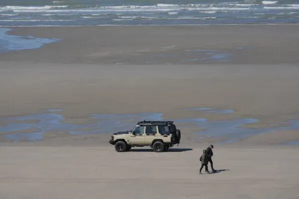 رجل يسير على شاطئ واسع بالقرب من مركبة شرطة، في إطار جهود مراقبة الهجرة غير النظامية عبر القناة الإنجليزية.