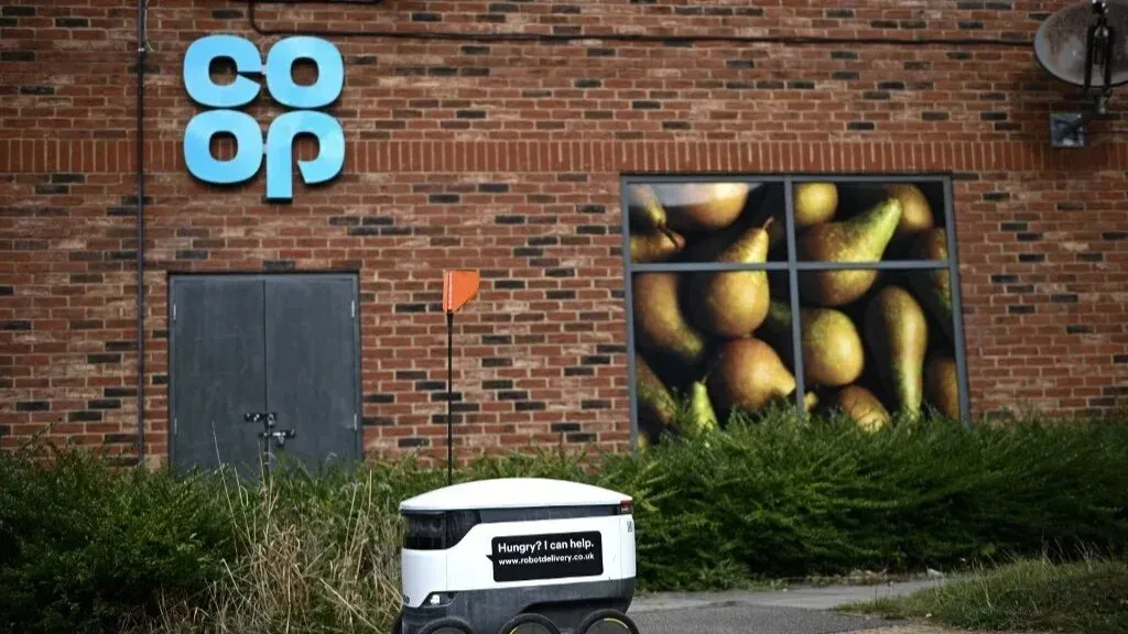 روبوت توصيل أمام واجهة متجر Co-operative Group، مع علامة المتجر على الجدار وصورة للخضروات في النافذة، يعكس جهود التوصيل الحديثة.