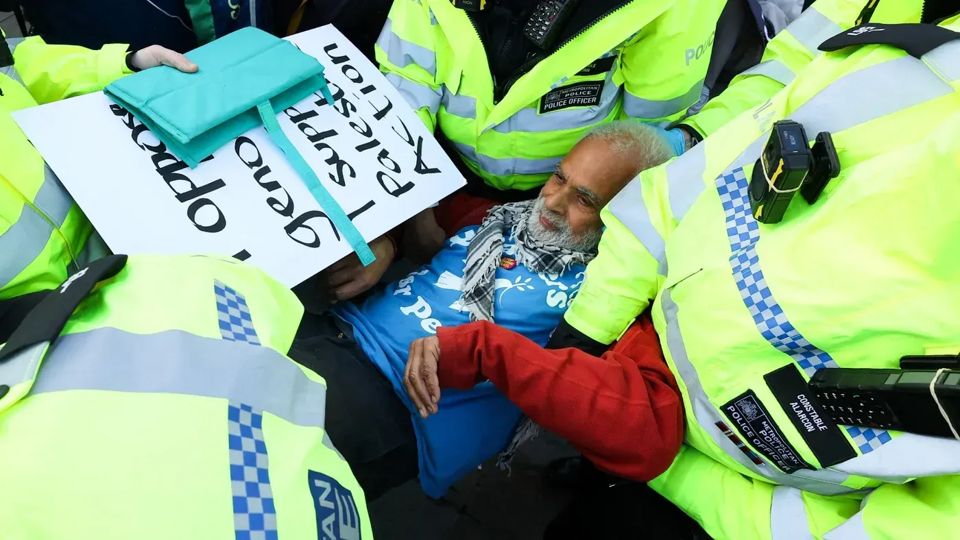 رجل مسن يرتدي قميصًا أزرق يحمل لافتة تدعم فلسطين، محاطًا برجال الشرطة خلال احتجاج ضد انتهاكات حقوق الإنسان.