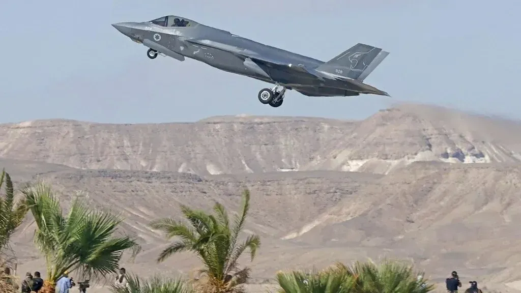 طائرة F-35 بريطانية الصنع تحلق فوق منطقة صحراوية، مع وجود أشخاص ومشاهد طبيعية في الخلفية، تعبيراً عن القلق بشأن تصدير الأسلحة.