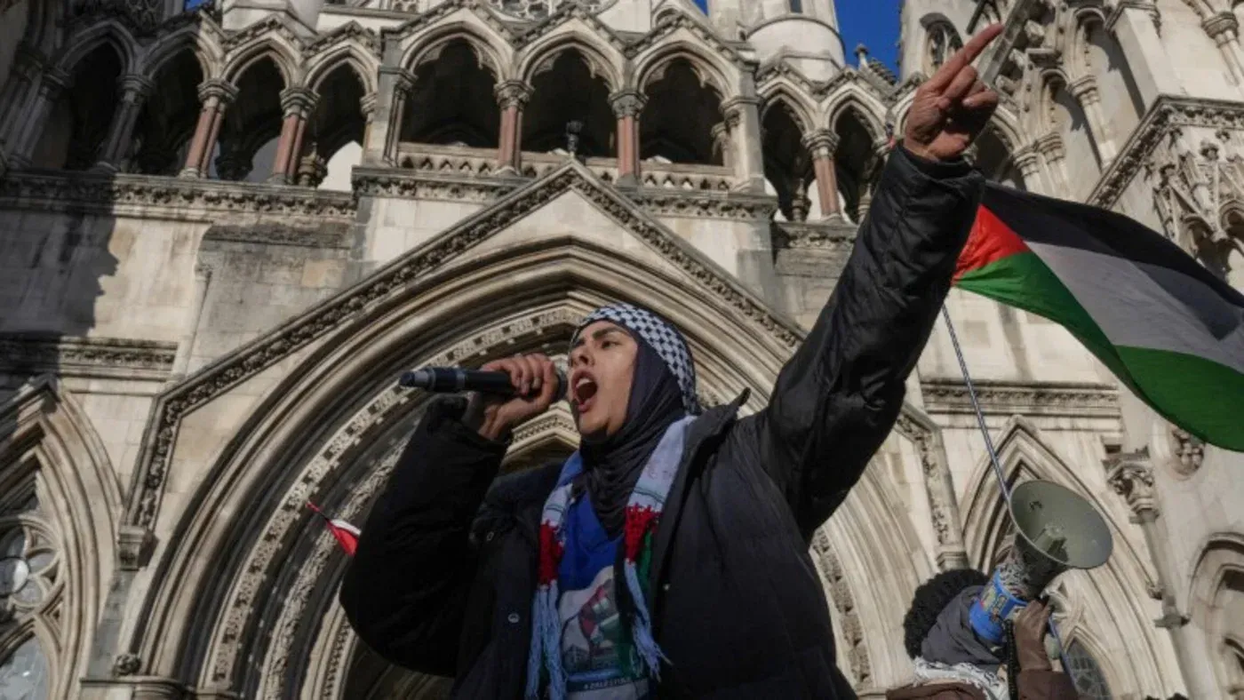 امرأة ترتدي حجابًا وتحتج أمام مبنى المحكمة، تحمل مكبر صوت وتلوح بعلم فلسطين، تعبر عن دعمها لمنظمة العمل الفلسطيني.