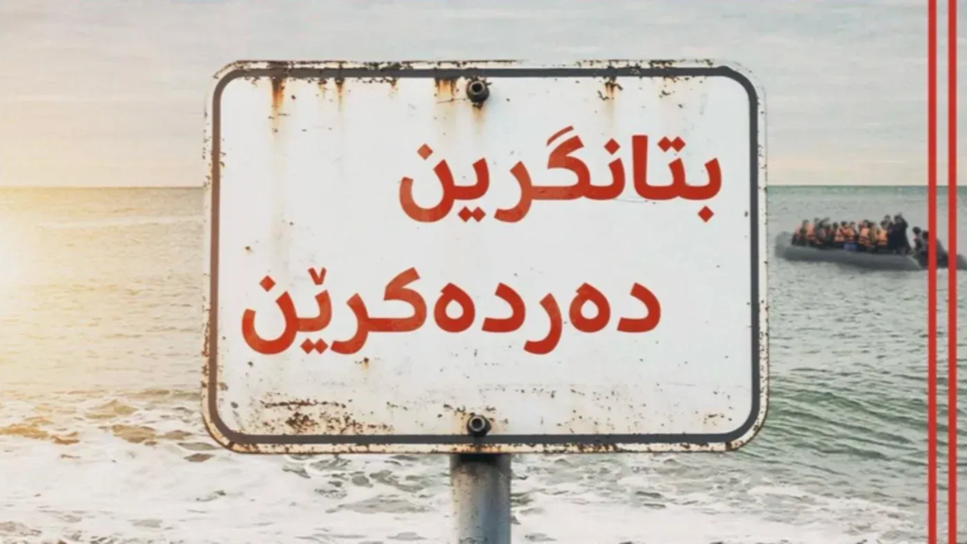 لافتة تحذيرية باللغة الكردية السورانية على شاطئ، تشير إلى مخاطر الهجرة غير الشرعية إلى المملكة المتحدة، مع قارب يحمل مهاجرين في الخلفية.