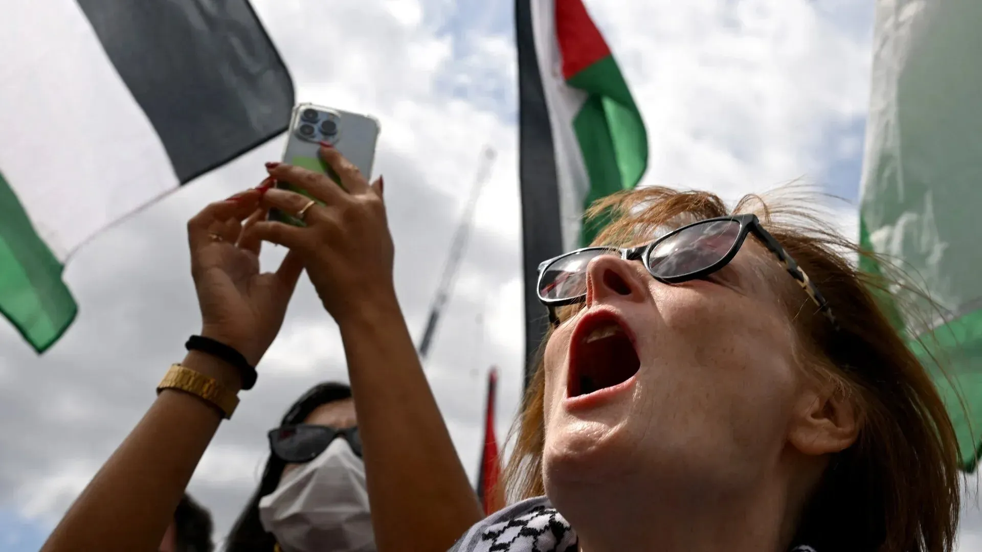 امرأة تتظاهر في احتجاج لدعم فلسطين، ترفع هاتفها وتصرخ بحماس، مع أعلام فلسطينية في الخلفية.
