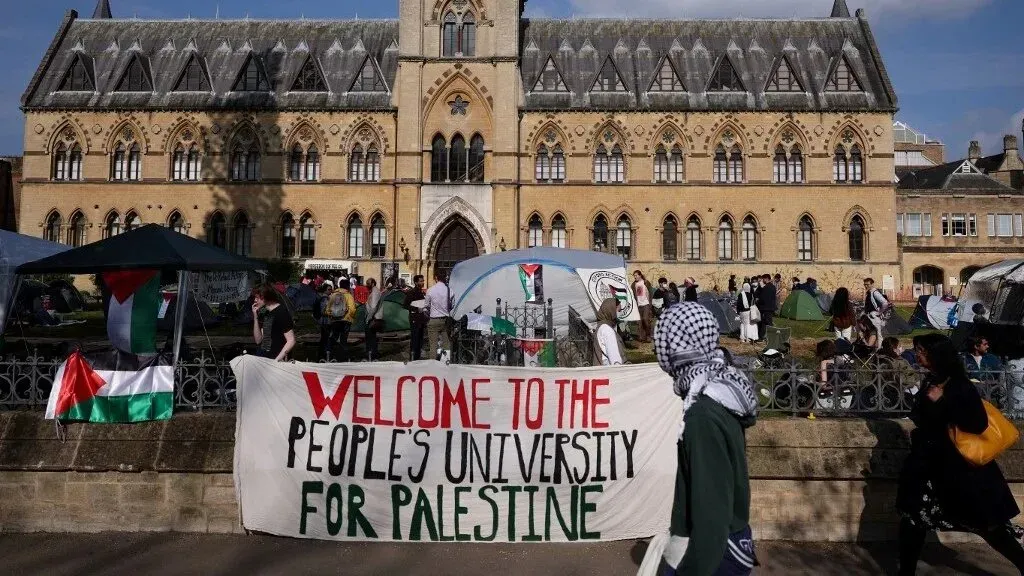 محتجون أمام مبنى تاريخي في أكسفورد يحملون لافتة ترحب بـ "جامعة الشعب لفلسطين"، مع أعلام فلسطينية وخيام.
