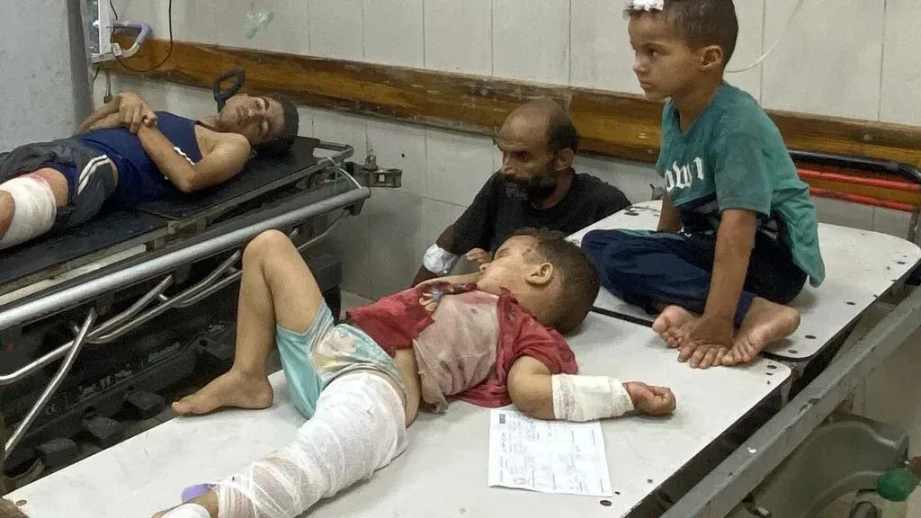 أطفال مصابون في مستشفى بغزة، يعانون من إصابات خطيرة، مع وجود بالغين بجوارهم، مما يعكس الحاجة الملحة للعلاج الطبي.