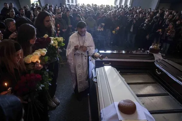 تجمع حشد كبير من الأشخاص في مراسم تأبين، حيث يقف كاهن أمام تابوت، محاطًا بالزهور والشموع، تعبيرًا عن الحزن لفقدان الأرواح في النزاع.