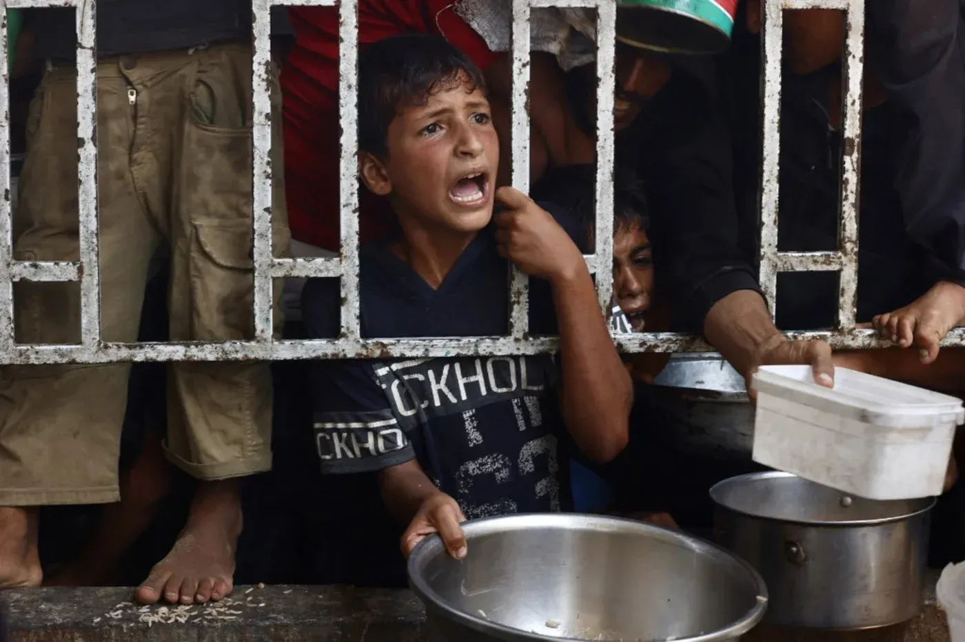 طفل فلسطيني يصرخ خلف قضبان حديدية أثناء محاولته الحصول على الطعام في مركز توزيع في غزة، وسط أزمة إنسانية حادة.