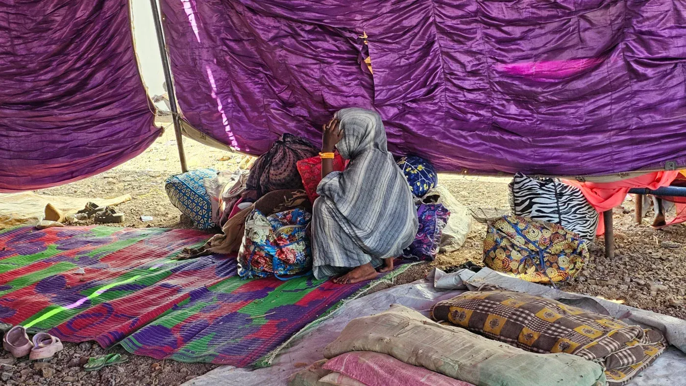 امرأة تجلس في مأوى مؤقت تحت خيمة، محاطة بأمتعتها، تعكس معاناة النازحين في الفاشر بالسودان بعد النزاع.